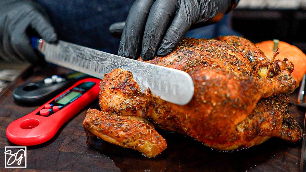A gloved hand slices a seasoned oven baked chicken breast on a cutting board with a patterned knife. A red digital meat thermometer is placed next to the chicken.