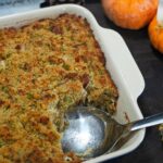 A baking dish filled with golden-brown cornbread dressing sits on a dark surface, with a serving spoon inside. Nearby are two small orange pumpkins and decorative ears of multicolored corn.