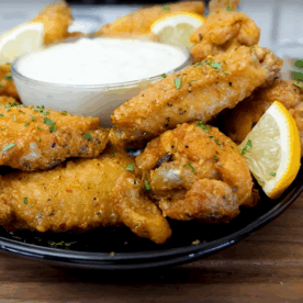 A black plate of crispy lemon pepper chicken wings garnished with lemon wedges and herbs, served with a bowl of creamy dipping sauce on a wooden table, with lemon slices in the background. Perfect for those craving a classic lemon pepper wings recipe.