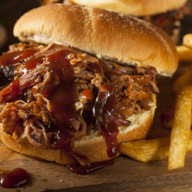 A classic BBQ pulled pork sandwich topped with smoky barbecue sauce, served on a wooden board alongside golden French fries.