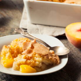A plate with a serving of peach cobbler, featuring golden crust and juicy peach filling, sits next to a spoon on a rustic wooden table. Nearby, Cajun Gravy adds a bold twist, while a halved peach and baking dish are visible in the background.