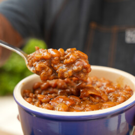 A person holds a blue bowl of Smoked BBQ Baked Beans Recipe in one hand and lifts a spoonful of the beans with the other. The beans are covered in a thick, rich sauce. The person is wearing a dark shirt and apron.