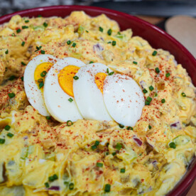 A bowl of creamy yellow deviled egg potato salad is garnished with four slices of hard-boiled egg, paprika, and chopped chives, placed on a wooden surface with a spoon nearby.