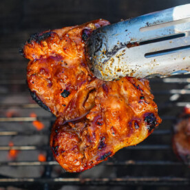 A close-up of metal tongs holding a piece of grilled, glazed huli huli chicken over a barbecue grill, with other pieces cooking in the background. Bright sunlight highlights the juicy texture and caramelized grill marks.