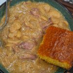 A bowl of creamy lima beans with chunks of meat is served with a square piece of golden cornbread. A spoon rests in the bowl, and a paper napkin is visible in the background.