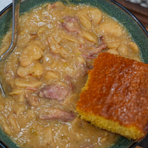 A bowl of creamy lima beans with chunks of meat is served with a square piece of golden cornbread. A spoon rests in the bowl, and a paper napkin is visible in the background.