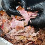 A person wearing black gloves is pulling apart juicy, smoked pork butt in an aluminum tray, revealing tender, shredded pieces—perfect inspiration if you're looking for smoked pork butt recipes.