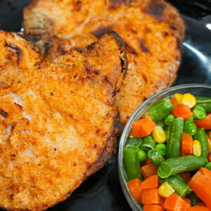 Two seasoned, grilled air fryer pork chops on a black plate sit next to a small bowl of mixed vegetables, including green beans, carrots, and corn. A fork and knife are partially visible beside the plate.