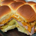 A close-up of golden, fluffy breakfast sliders stuffed with melted cheddar cheese, sliced ham, and a folded omelette in a clear baking dish—a delicious breakfast sliders recipe everyone will love.