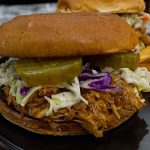 A pulled BBQ chicken sandwich crockpot-style, topped with coleslaw and sliced pickles on a toasted bun, is served on a black plate next to a stack of ridged potato chips.