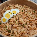 A wooden bowl filled with creamy macaroni salad, garnished with chopped chives and topped with slices of hard-boiled egg, sits on a kitchen counter—an inviting sight for anyone craving a classic macaroni salad recipe.