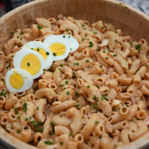 A wooden bowl filled with creamy macaroni salad, garnished with chopped chives and topped with slices of hard-boiled egg, sits on a kitchen counter&mdash;an inviting sight for anyone craving a classic macaroni salad recipe.