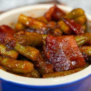 A blue-rimmed bowl filled with stewed green beans and chunks of bacon in a rich, dark sauce is a comforting twist on the classic green bean recipe, placed invitingly on a wooden surface.
