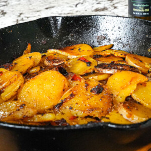 Sliced, seasoned potatoes and onions cook in a black cast iron skillet, appearing golden brown and crispy. A bottle of avocado oil and seasoning are visible in the background, perfect for a classic roasted potatoes and onions recipe.