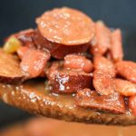A wooden spoon holds a serving of red beans and sliced sausage in a thick, savory sauce, perfect for anyone seeking classic red beans and rice recipes, with a blurred background of a pot.