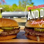 Two cheeseburgers with pickles sit on a wooden board outdoors, a grill in the background. Text overlays the image: Burgers and Dogs Across America in bold white and red letters, featuring an Oklahoma fried onion burger.