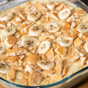 A glass dish filled with banana pudding, topped with sliced bananas and crumbled vanilla wafer cookies, sits on a wooden surface&mdash;perfect for anyone searching for an easy banana pudding recipe.