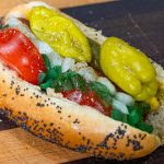A classic Chicago-style hot dog in a poppy seed bun topped with tomatoes, onions, sport peppers, pickles, relish, mustard, and celery salt is served on a wooden surface. This authentic Chicago dog bursts with flavor in every bite.