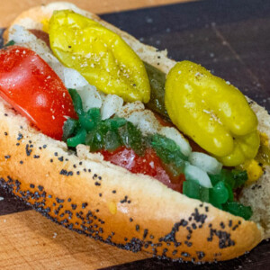 A classic Chicago-style hot dog in a poppy seed bun topped with tomatoes, onions, sport peppers, pickles, relish, mustard, and celery salt is served on a wooden surface. This authentic Chicago dog bursts with flavor in every bite.