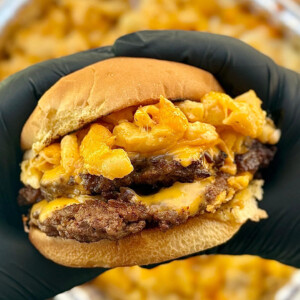 A person wearing black gloves holds a cheeseburger filled with two beef patties, melted cheese, and creamy smoked gouda mac and cheese, with a tray of smoked mac and cheese in the background.