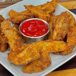 A square white plate holds a serving of crispy, golden-brown air fryer potato wedges surrounding a small metal cup filled with red ketchup, set on a striped wooden surface.