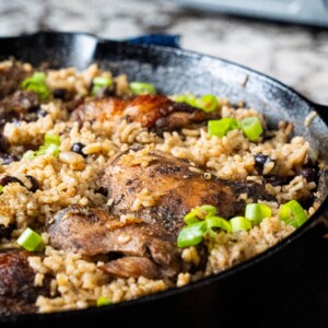 A close-up of a cast iron skillet filled with seasoned rice, black beans, and pieces of browned jerk chicken, garnished with chopped green onions.