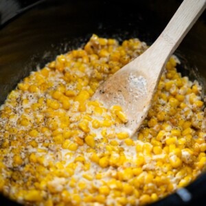 Creamed corn being mixed with cream and seasonings in a slow cooker, stirred gently with a wooden spoon&mdash;this classic creamed corn recipe is perfect for cozy meals and family gatherings.