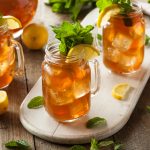 Three mason jars of southern sweet tea with lemon slices and fresh mint on a wooden tray, surrounded by lemon halves and mint leaves on a rustic wooden table.