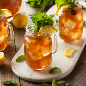 Three mason jars of southern sweet tea with lemon slices and fresh mint on a wooden tray, surrounded by lemon halves and mint leaves on a rustic wooden table.