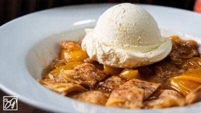 A close-up of a white bowl filled with easy peach cobbler, topped with a scoop of vanilla ice cream. The cobbler features golden crust pieces and juicy peach slices, perfect for fans of classic peach cobbler recipes.