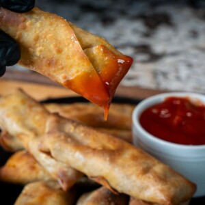 A gloved hand holds a crispy cheeseburger eggroll dipped in red sauce, above a plate stacked with more cheeseburger eggrolls and a small white bowl of sauce on the side.