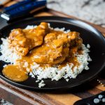 A black plate with white rice topped with pieces of smothered chicken and gravy, placed on a wooden cutting board. Knives and a digital thermometer are nearby.