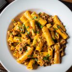 A white bowl filled with creamy cajun pasta, rigatoni in a savory meat sauce and garnished with chopped herbs, sits on a wooden surface next to a large kitchen knife.
