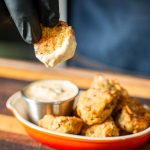 A gloved hand dips a crispy, golden fried pickle into a cup of creamy sauce, with more fried pickles piled in an orange dish on a wooden surface nearby.