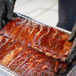 A person wearing black gloves is placing two racks of smoked ribs into a metal tray, showing the final step in how to smoke ribs before serving.