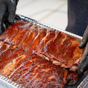 A person wearing black gloves is placing two racks of smoked ribs into a metal tray, showing the final step in how to smoke ribs before serving.