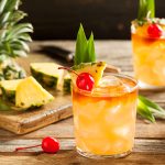 Three tropical cocktails with ice, garnished with pineapple wedges, cherries, and leaves, sit on a wooden table—perfect for trying a classic mai tai recipe. In the background are pineapple slices on a cutting board and a whole pineapple.