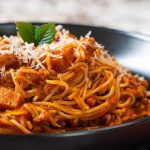 A close-up of a plate of spaghetti with tomato-based meat sauce, topped with grated cheese and fresh basil, served in a black bowl—perfect inspiration for your next spaghetti sauce recipe.