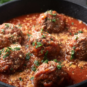 Six meatballs, made from a classic meatball recipe, are topped with grated cheese and chopped parsley in a cast iron skillet filled with rich tomato sauce.