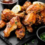 A pile of crispy oven baked chicken wings glazed to perfection, garnished with chopped herbs and lemon slices, served on a black slate plate with dipping sauces and a small bowl of fresh parsley on the side.