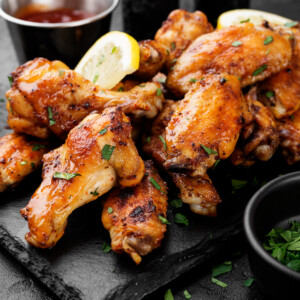 A pile of crispy oven baked chicken wings glazed to perfection, garnished with chopped herbs and lemon slices, served on a black slate plate with dipping sauces and a small bowl of fresh parsley on the side.