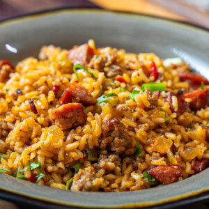 A bowl of Cajun dirty rice mixed with sausage, ground meat, chopped green onions, and herbs, served in a rustic gray dish on a wooden table.