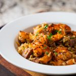 A white bowl filled with shrimp and rice, garnished with herbs, sits on a wooden cutting board next to a fork; this shrimp and rice recipe appears flavorful and freshly prepared.