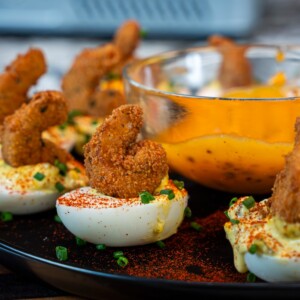 Deviled Eggs topped with fried shrimp and chopped chives are arranged on a black platter, with a glass bowl of orange dipping sauce in the center&mdash;a unique twist on the classic deviled egg recipe.