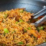 A close-up of a pan filled with stir-fried chicken ramen noodles, chopped green onions, broccoli, and carrots, with a pair of metal tongs resting in the pan—perfect for trying new chicken ramen noodle recipes.