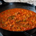 A cast iron skillet filled with chunky, tomato-based stew, featuring stewed okra and tomatoes with visible pieces of vegetables, sits on a stove against a marble countertop background.