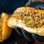 A hand wearing a black glove holds a beef and cheese sandwich filled with creamy, cheesy steak and chopped green herbs in a soft, toasted bun. Another similar sandwich is blurred in the background.