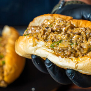 A hand wearing a black glove holds a beef and cheese sandwich filled with creamy, cheesy steak and chopped green herbs in a soft, toasted bun. Another similar sandwich is blurred in the background.