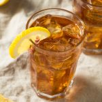 A glass of southern style Gold Peak Sweet Tea with ice cubes, garnished with a lemon wedge and a striped straw, sits on a light-colored cloth with lemon slices nearby.