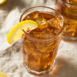 A glass of southern style Gold Peak Sweet Tea with ice cubes, garnished with a lemon wedge and a striped straw, sits on a light-colored cloth with lemon slices nearby.